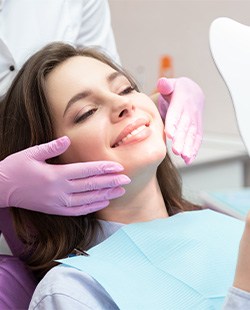 Woman smiling into handheld mirror sitting in dental chair